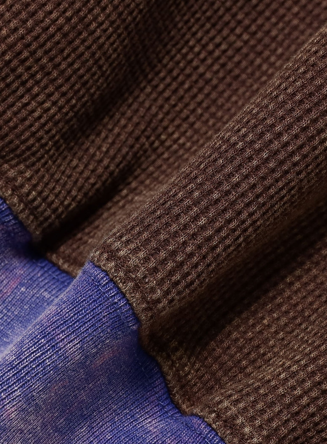 Close-up of textured brown waffle knit fabric overlapping a purple-blue denim-like material, with lighting that accentuates their contrast and detail—evoking the vintage-inspired design of the TTT MSW Fade Damage Zip Up Hoodie.