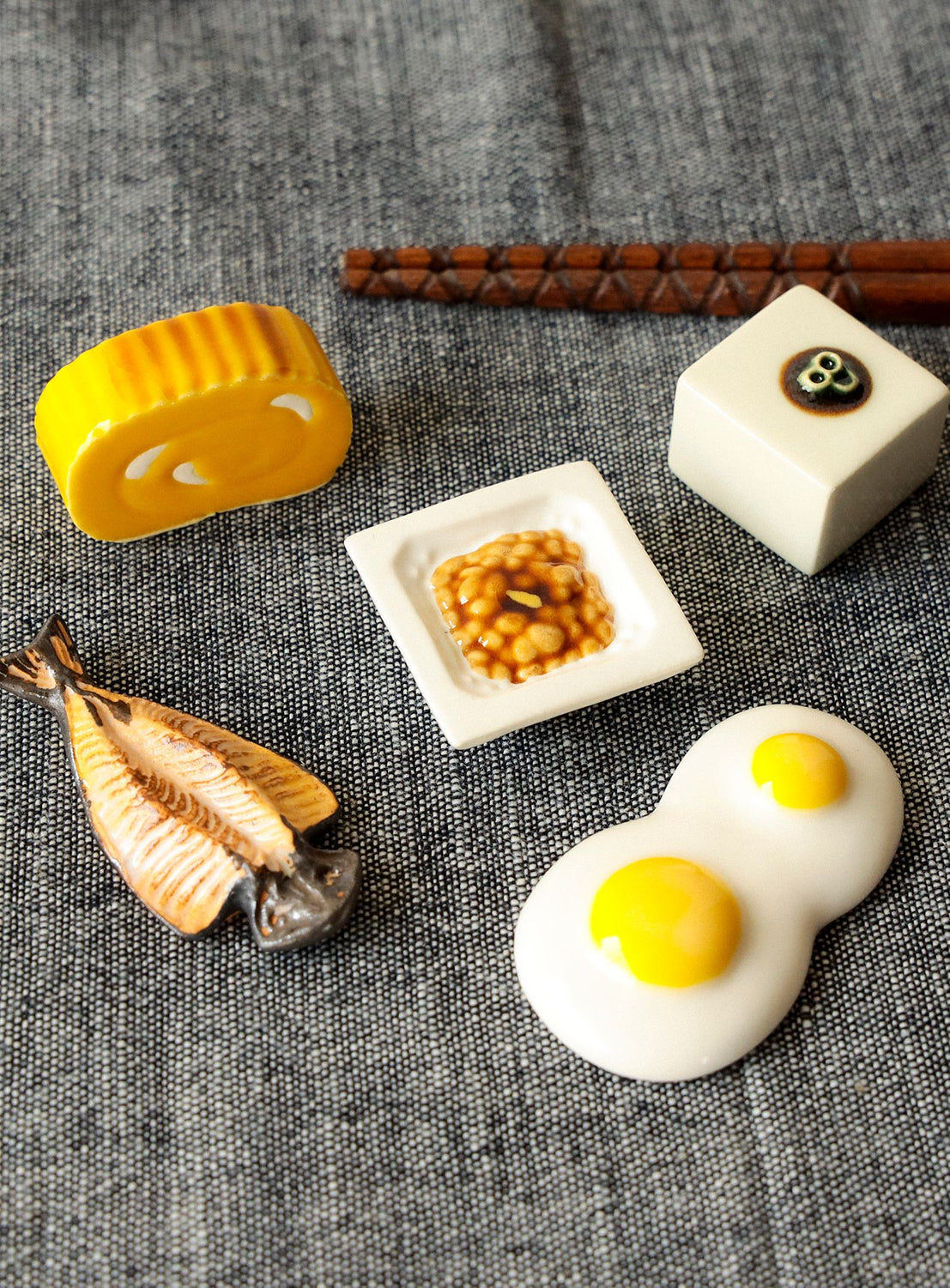 Miniature food item chopstick rests including a yellow block, white square with a brown center, and a white egg with a yellow yolk on a textured gray surface.