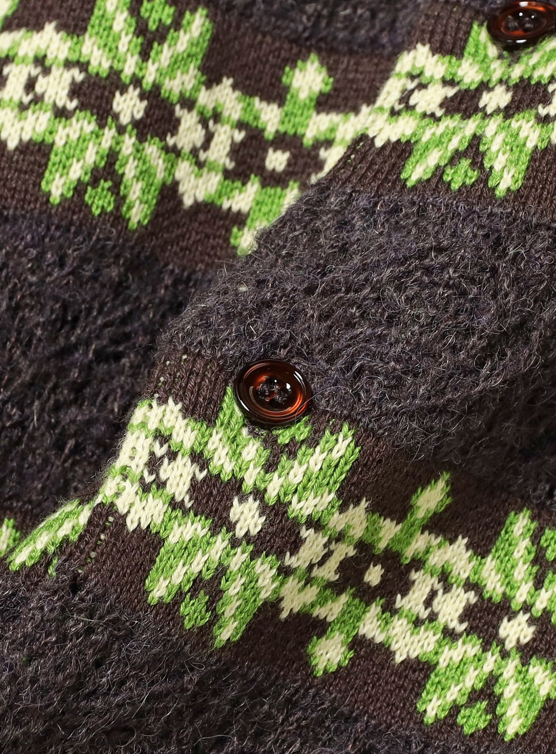 Close-up of the TTT MSW Crewneck Cardigan, showcasing green and white snowflake jacquard patterns on a dark brown background and finished with a brown button.