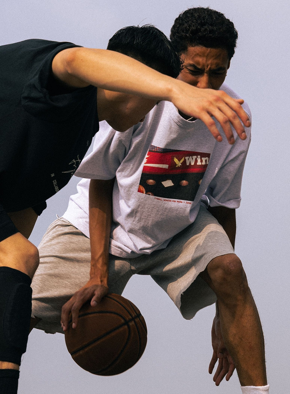 Two young people play basketball outdoors, both radiating dynamic energy reminiscent of a BEAMS T moment. One dribbles the ball while the other closely defends, both focused and active—one sporting the Tyrrell Winston - Win T-Shirt from BEAMS T against a backdrop of clear skies.