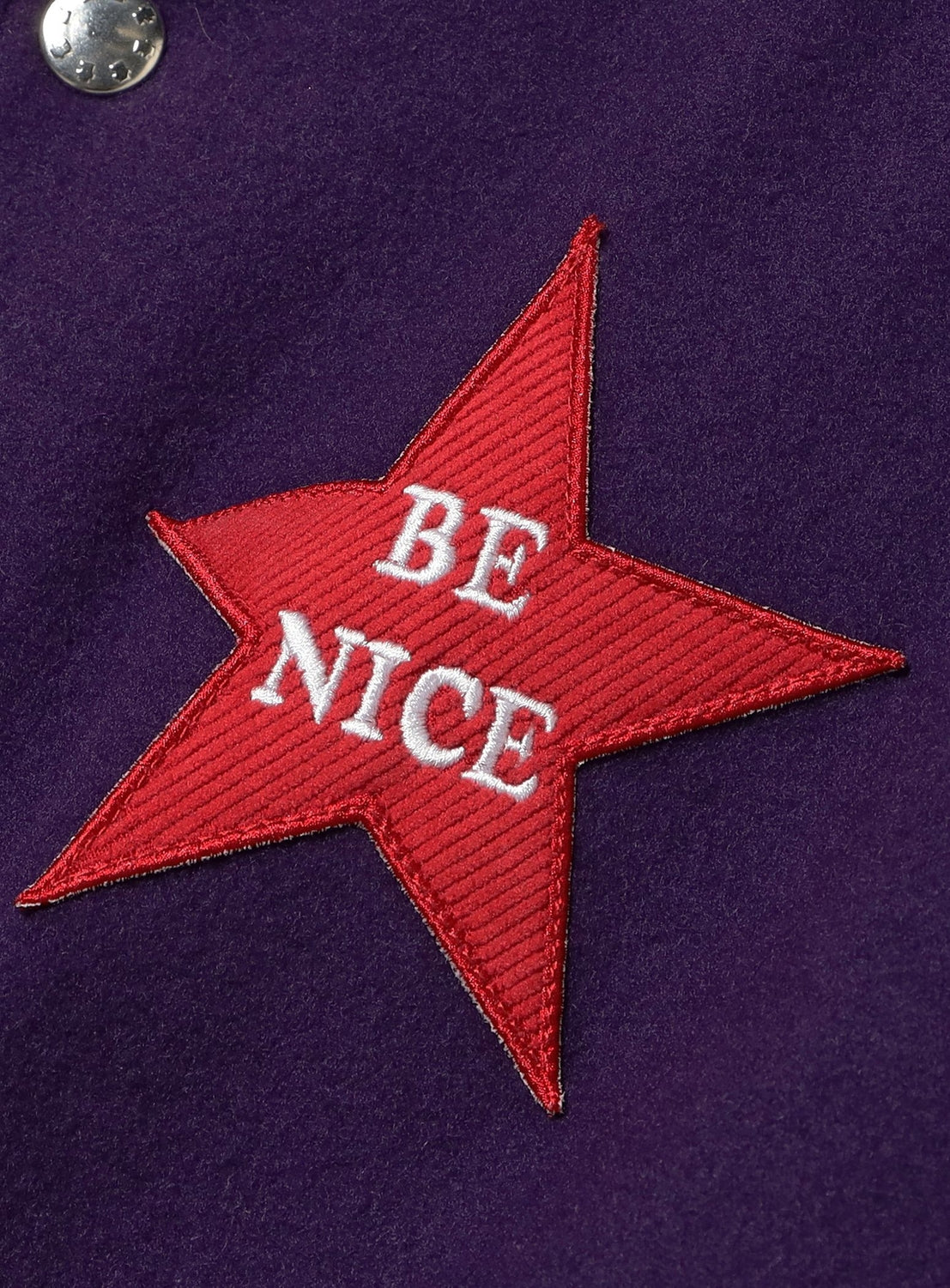 The TTT MSW Varsity Patch Jacket features a red, star-shaped patch with "BE NICE" in white lettering sewn onto its dark purple lamb wool fabric, bringing a playful accent to this modern-fit classic.