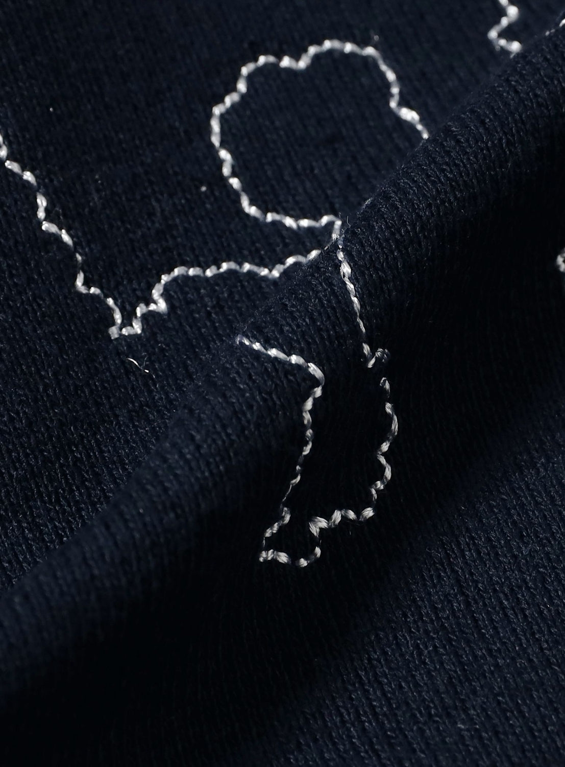 Close-up of the dark blue textured fabric from the TTT MSW Poodle Embroidery Zip-Up Hoodie, featuring white, irregular wavy stitched lines that create abstract patterns. The slightly folded fabric emphasizes its intricate texture and detailed embroidery.