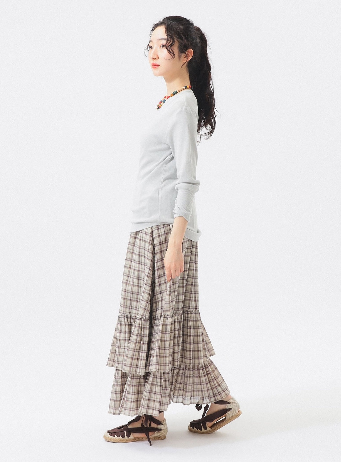 A woman stands sideways against a plain white background, her hair tied back in a loose ponytail. She is wearing a light long-sleeve top paired with the BEAMS BOY Check Tiered Skirt made from lightweight fabric. Her look is completed with brown sandals, glasses, and a colorful beaded necklace.