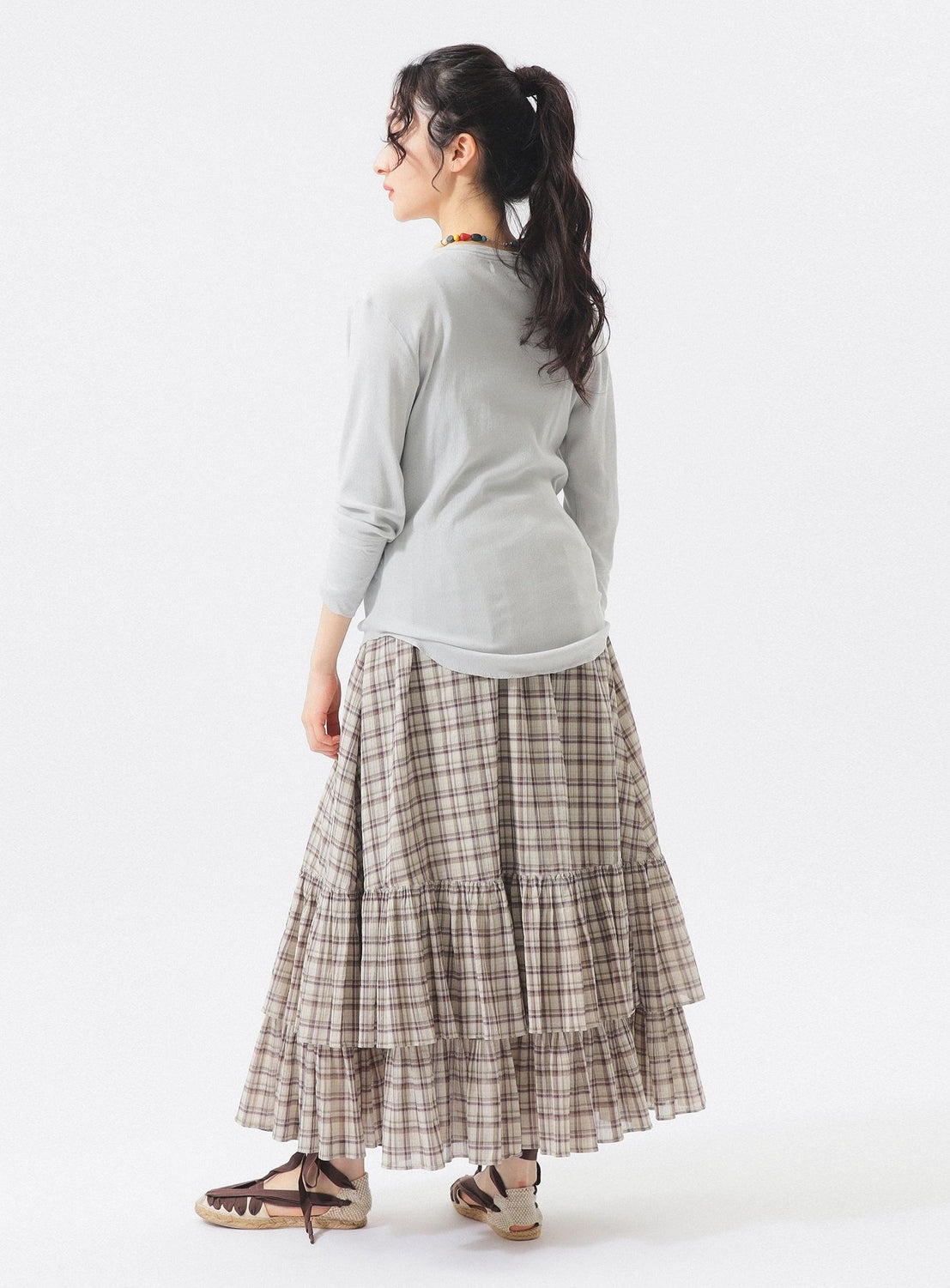 A woman stands facing away, wearing a light long-sleeve top paired with the BEAMS BOY Check Tiered Skirt, featuring a brown plaid pattern crafted from lightweight fabric. She completes the look with brown shoes and has her long dark hair tied in a ponytail. The distinctive check pattern of the BEAMS BOY Check Tiered Skirt stands out against the plain white background.