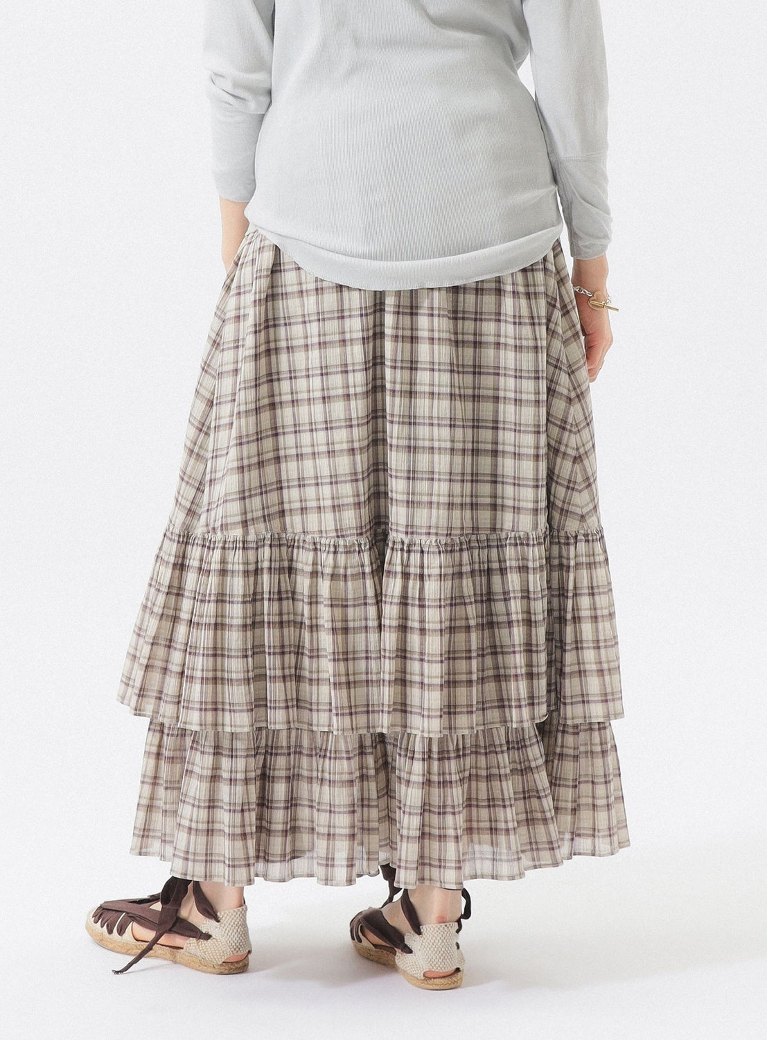 A person stands facing away on a white background, wearing a light long-sleeve shirt, the BEAMS BOY Check Tiered Skirt crafted from lightweight fabric with a signature check pattern, and espadrille sandals.