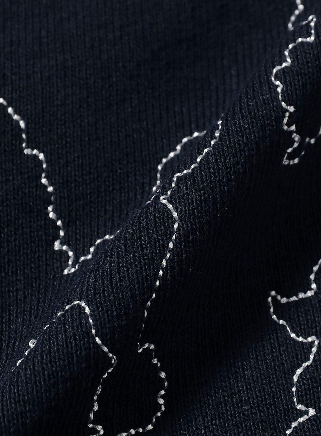 A close-up of dark textured fabric featuring white, irregular stitched lines that create abstract shapes and curves, similar to the detailed embroidery seen on TTT MSW's Dog Embroidery Half Pants.