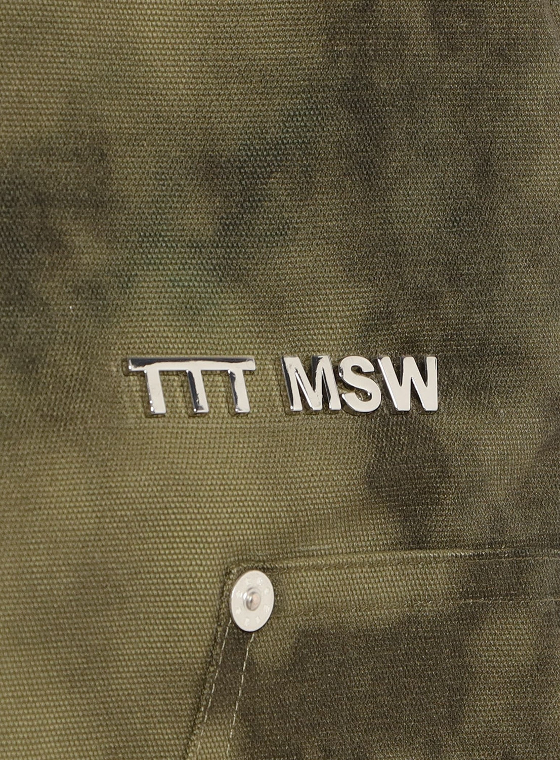 A close-up of the olive green heavy cotton twill fabric of the TTT MSW Camouflage Double Knee Pants, featuring a silver TTT MSW pin and a button visible near the bottom.