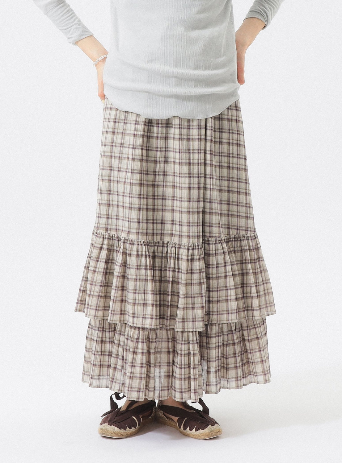 A person stands against a white background with hands on hips, wearing the BEAMS BOY Check Tiered Skirt—crafted from lightweight fabric in a signature plaid pattern and adorned with ruffles—paired with a light gray long-sleeve top and dark brown shoes featuring light-colored laces.