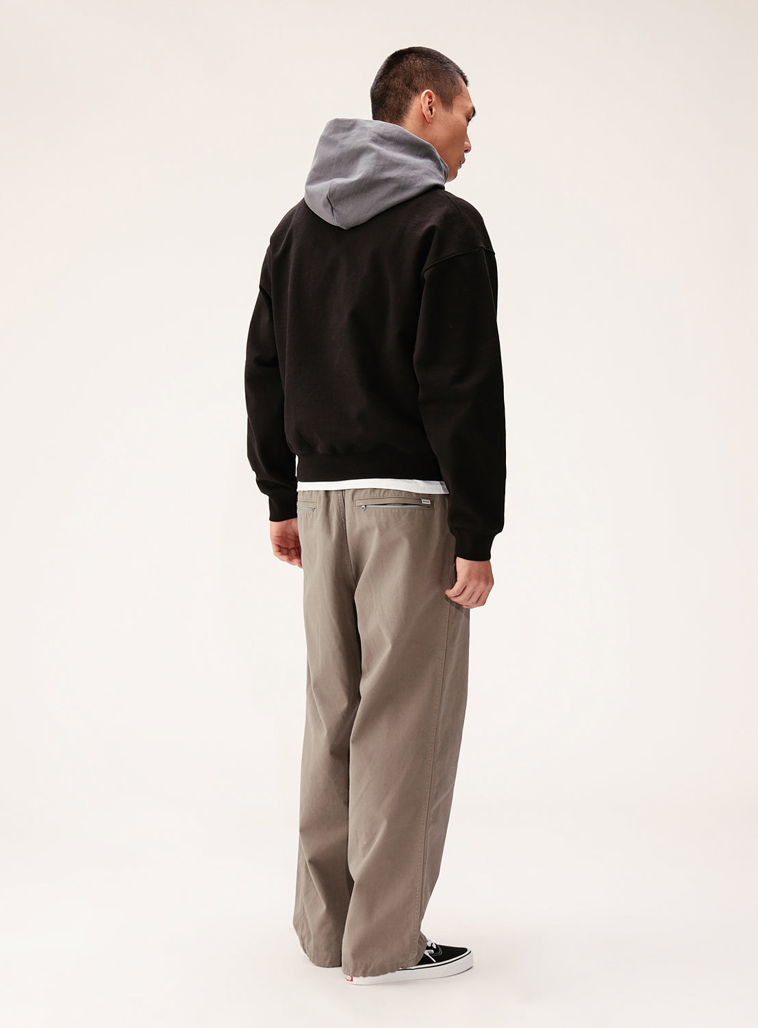 A person faces away, wearing the BEAMS Color Block Heavyweight Fleece Hoodie with a gray hood, paired with beige wide-leg pants and black slip-on shoes, against a plain light background.
