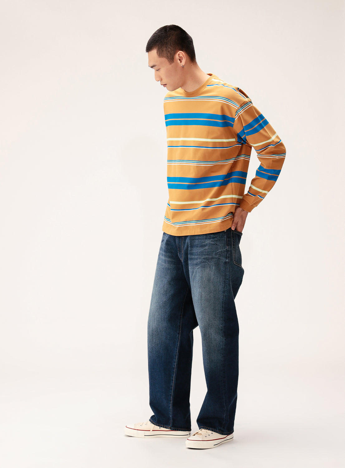 A man stands against a light background, looking down with his hands behind his back. He wears a BEAMS Multi-Stripe Crew Neck Wide Fit Sweatshirt in yellow, blue, and white, loose-fit blue jeans, and white sneakers.
