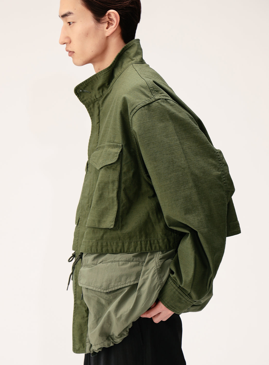 A person in profile wears the BEAMS Wide Fit M-65 Layered Jacket, featuring a two-tone green military back satin design with large front pockets, a high collar, and a relaxed, adjustable silhouette. The jacket is paired with dark pants against a light background.