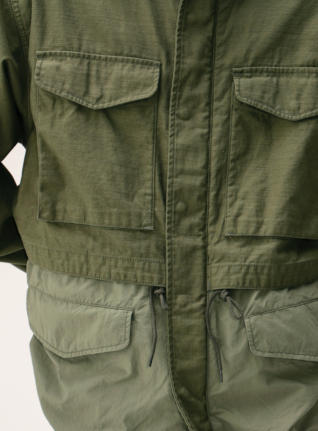 Close-up of the BEAMS Wide Fit M-65 Layered Jacket in olive green, featuring two flap chest pockets, a drawstring waist for an adjustable silhouette, visible stitching detail, and contrasting fabric textures on the upper and lower sections.