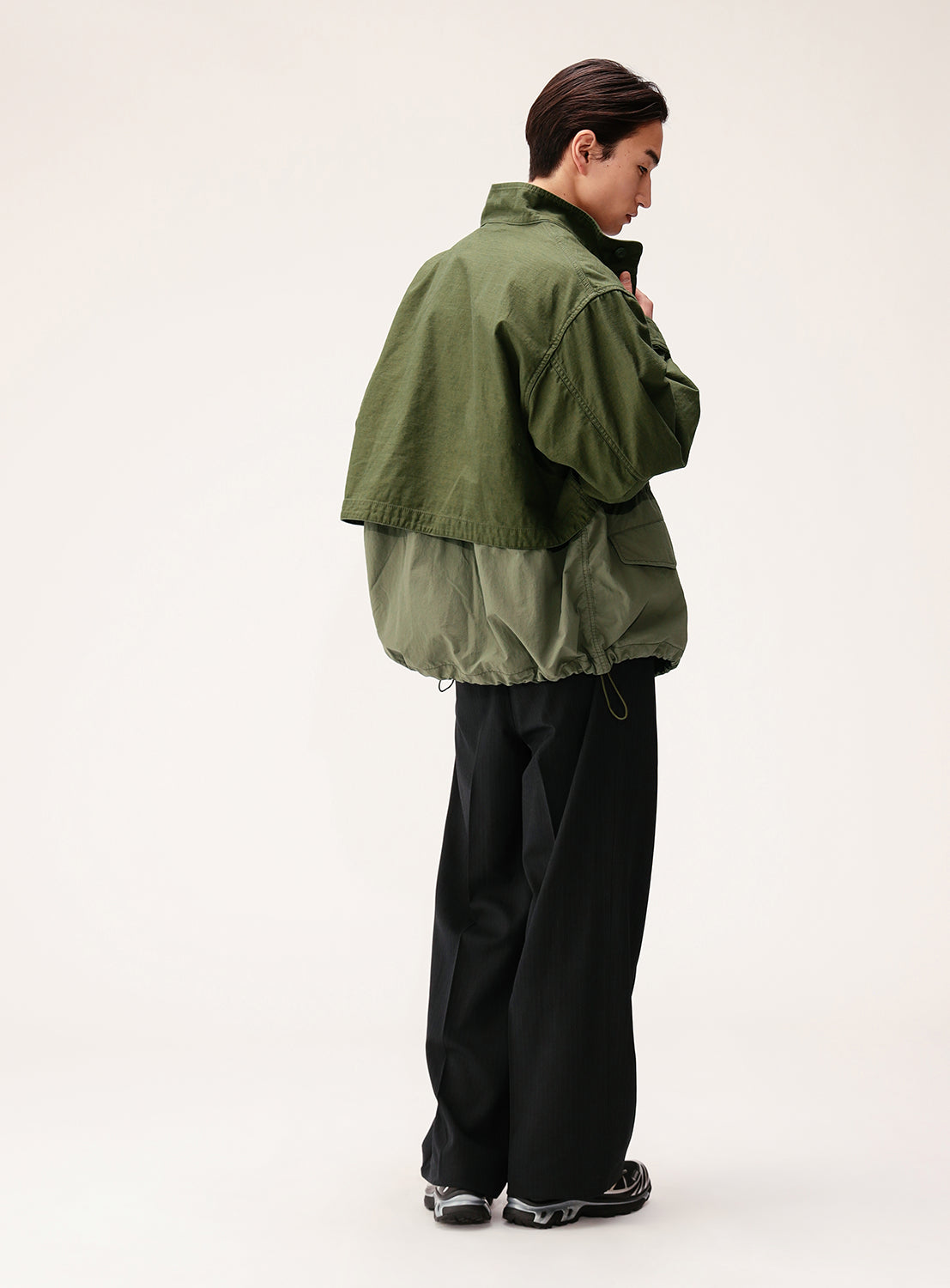 Facing away from the camera, the person is styled in BEAMS’ Wide Fit M-65 Layered Jacket, featuring a two-tone green back satin with a relaxed, adjustable silhouette. Paired with black wide-leg pants, the ensemble highlights their short dark hair and stands out against a plain light background.