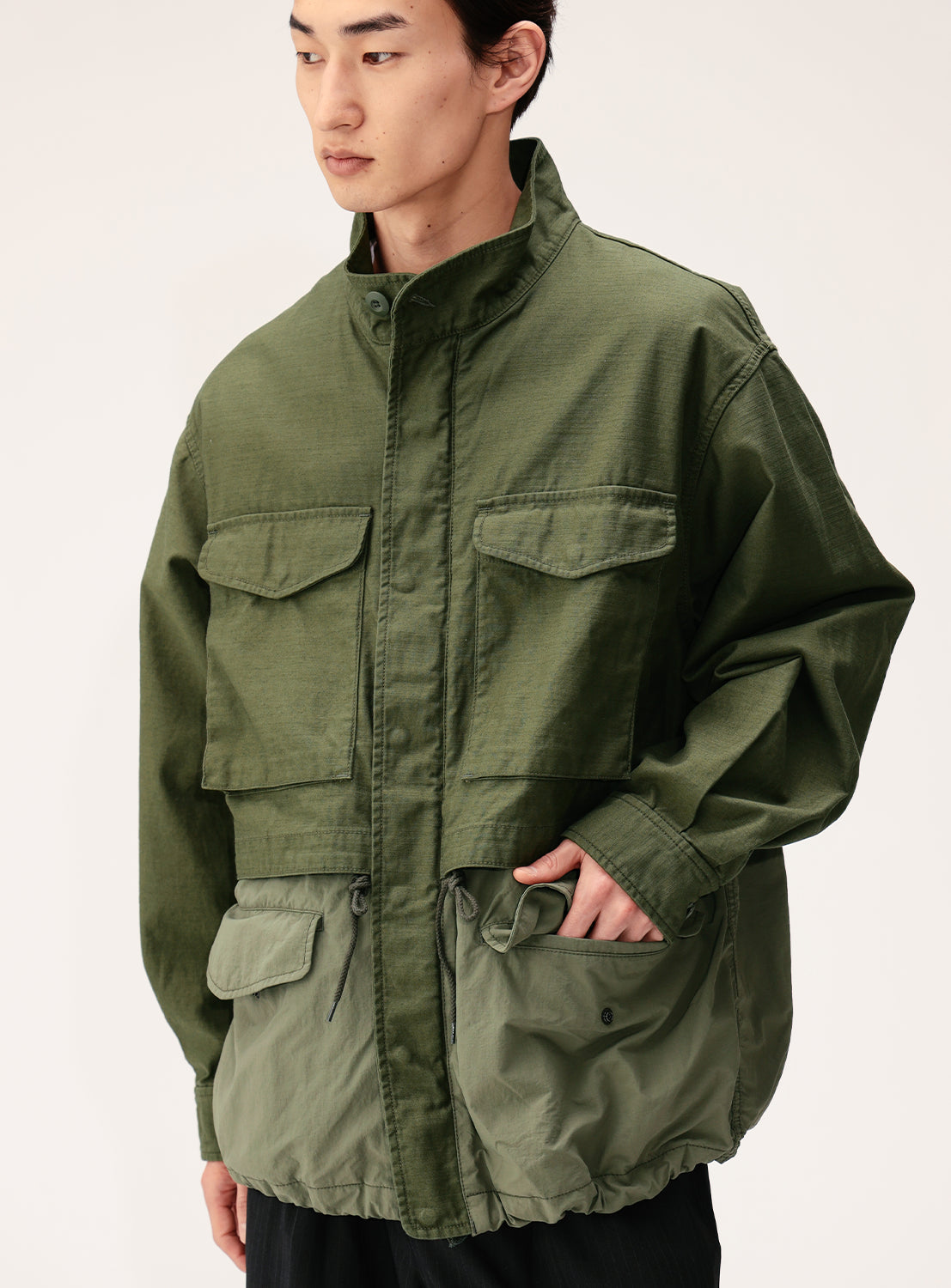 A person wearing the BEAMS Wide Fit M-65 Layered Jacket in olive green, featuring a loose fit, large front pockets, high collar, and an adjustable silhouette, stands against a plain light background.