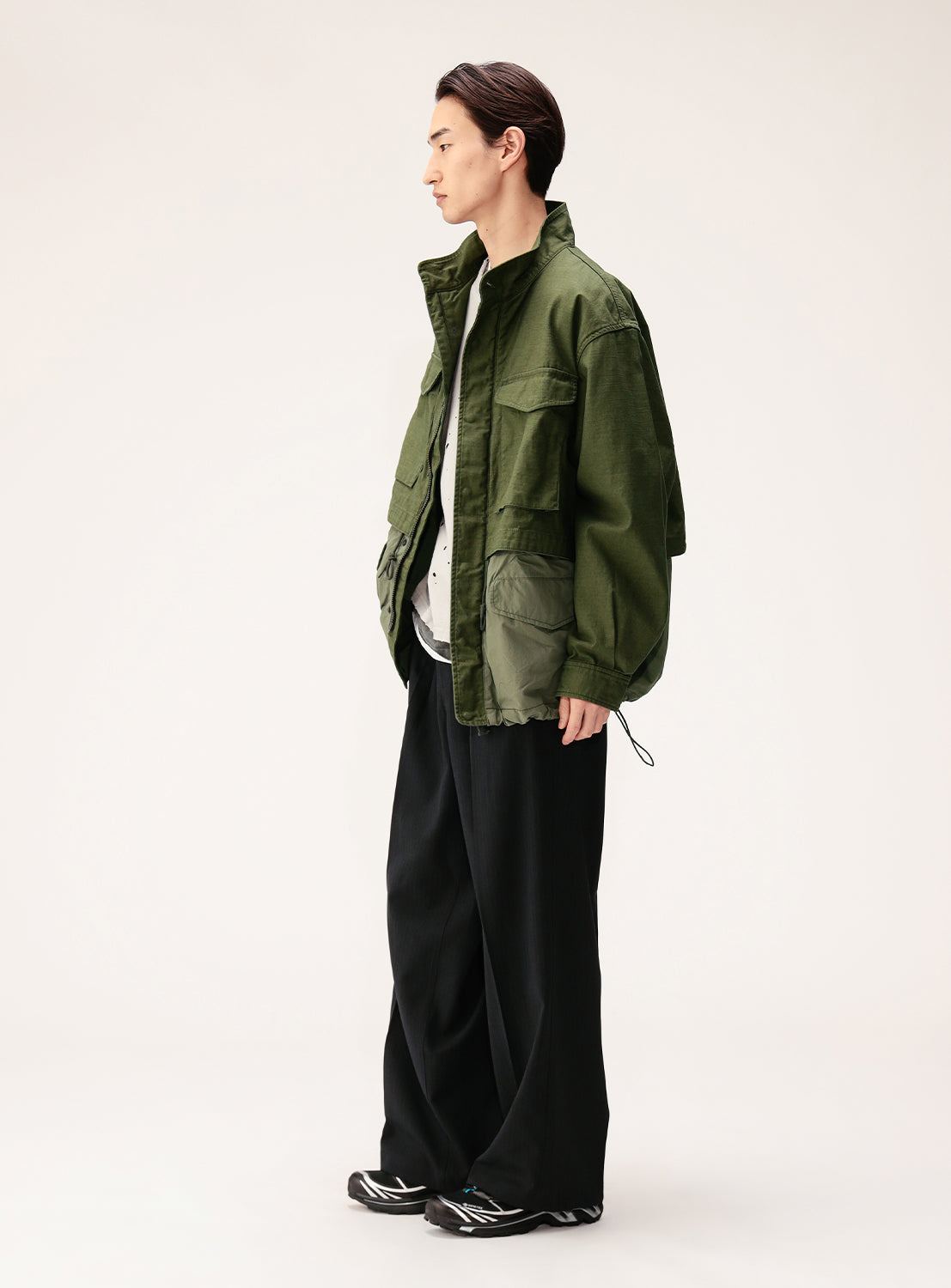 A person stands sideways against a plain light background, wearing the BEAMS Wide Fit M-65 Layered Jacket in green back satin with a double-zip front, layered over a white shirt, and styled with wide black pants and black-and-white sneakers.