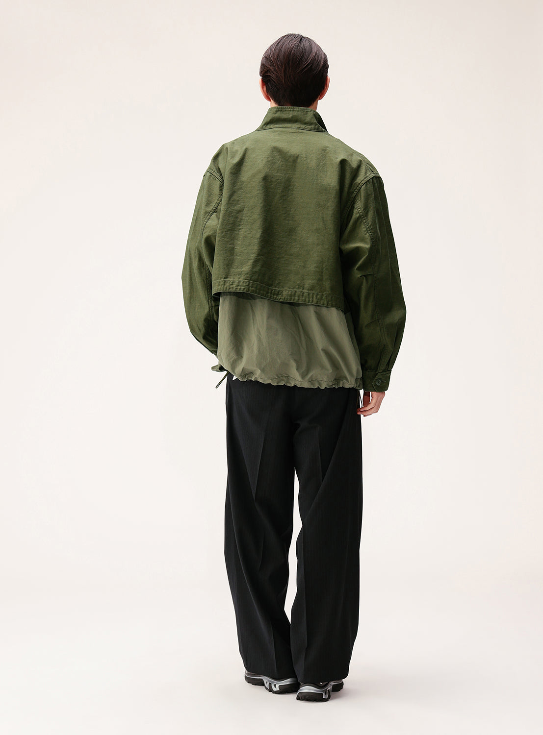 A person stands with their back to the camera, wearing the BEAMS Wide Fit M-65 Layered Jacket in two-tone green with an adjustable silhouette, paired with black pants, set against a plain light background.