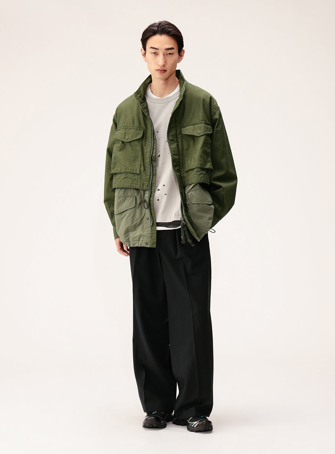 A person stands against a plain background wearing the BEAMS Wide Fit M-65 Layered Jacket in green with an adjustable silhouette over a white t-shirt, loose black pants, and black shoes, hands tucked into the jacket’s pockets.