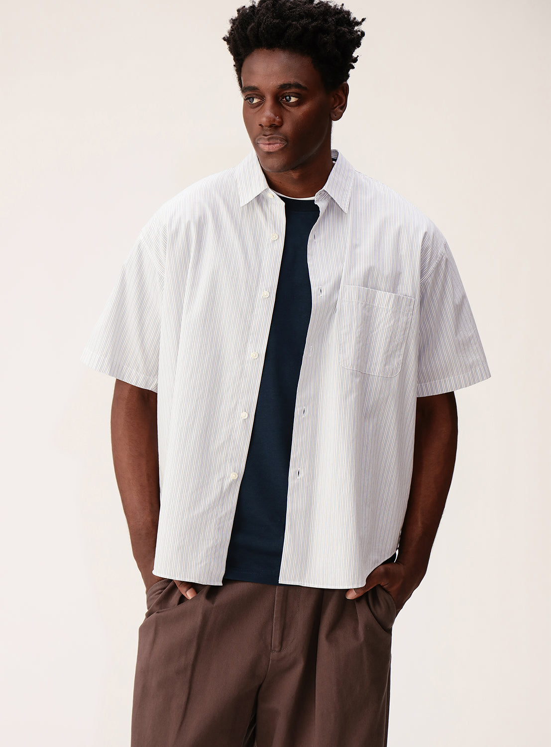 A man stands with his hands in his pockets, wearing the BEAMS Relaxed Fit Striped Shirt—a light blue, short-sleeve button-up—layered over a navy T-shirt and paired with brown pants, set against a plain light background.