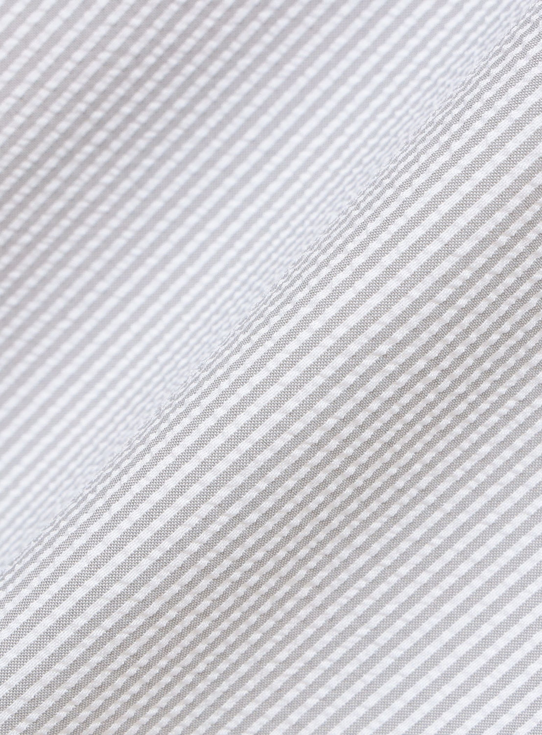Close-up of the BEAMS Stretch Quick-Dry Seersucker Relaxed Fit Shirt fabric, featuring light gray and white narrow stripes running diagonally across the image. The angled view highlights the subtle textured pattern and showcases the lightweight, wrinkle-resistant qualities of this seersucker material.