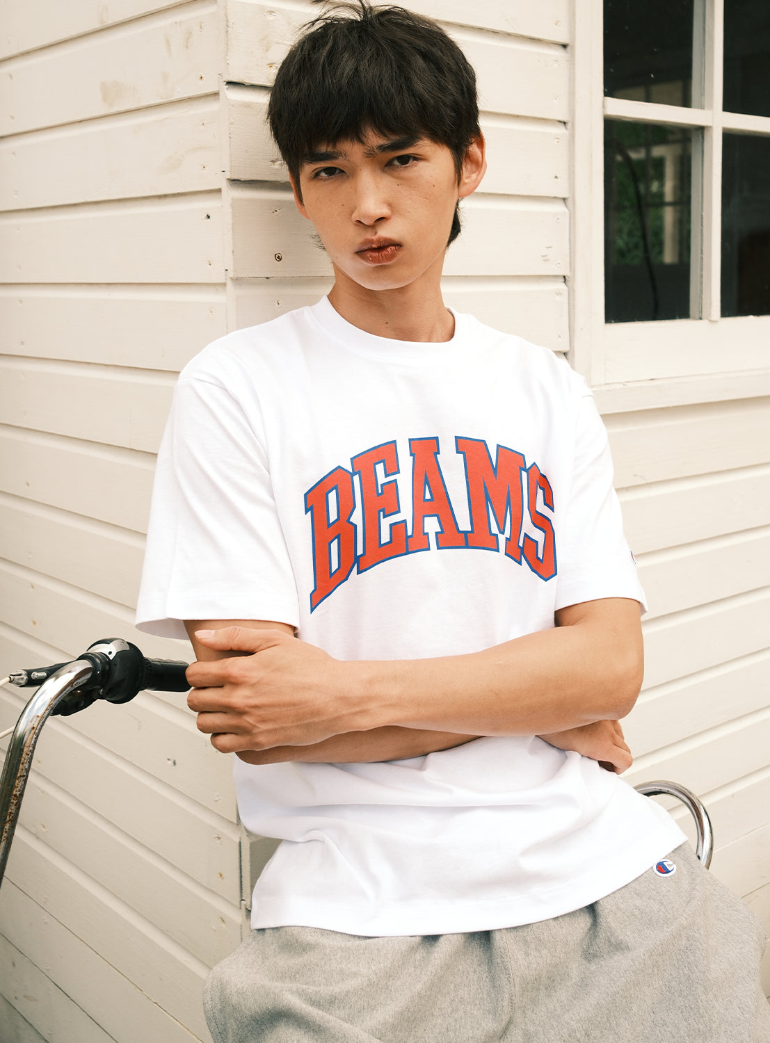 A person with short dark hair wears a white [Exclusive] BEAMS x Champion Logo T-Shirt by CHAMPION and gray pants, sitting on a bicycle in front of a white wooden building with arms crossed.