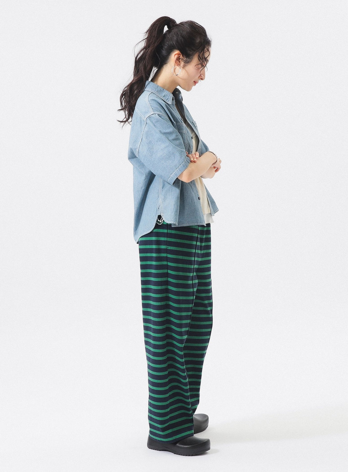 A woman in profile with arms crossed wears a BEAMS BOY Chambray Short-Sleeve Work Shirt over a white top, paired with loose green and black striped pants and black shoes. Her dark hair is in a high ponytail against a plain white background.