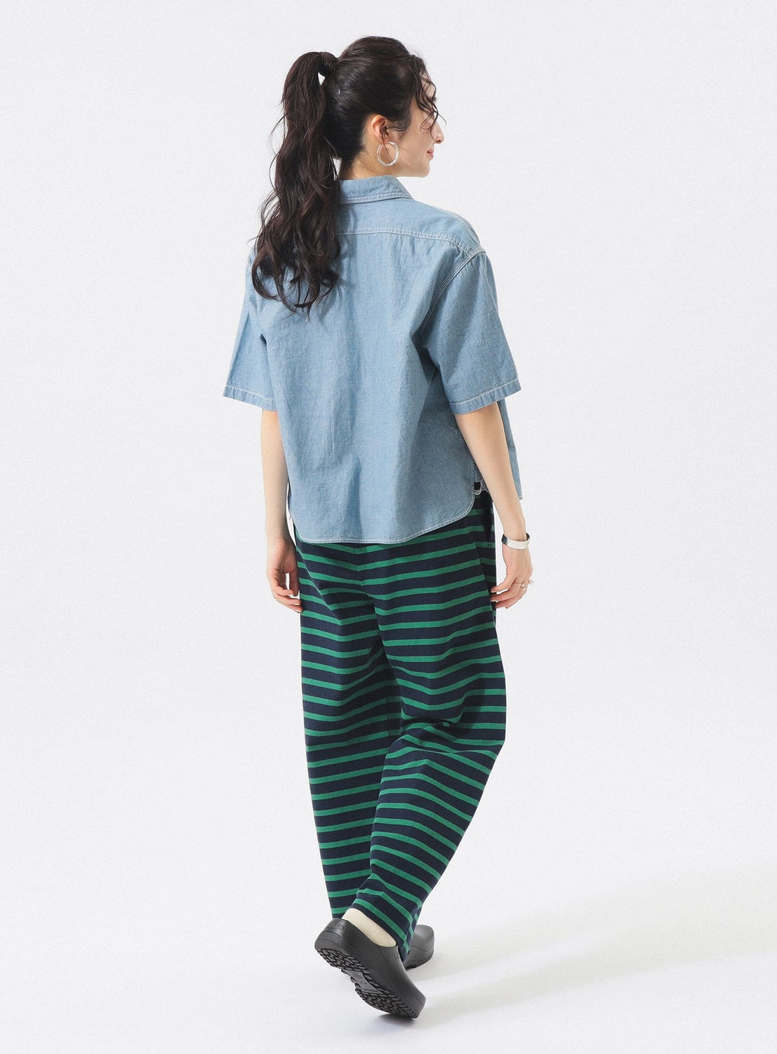 A person with long dark hair in a ponytail walks away against a plain white background, wearing a BEAMS BOY Chambray Short-Sleeve Work Shirt and loose green-and-black striped pants with black shoes.