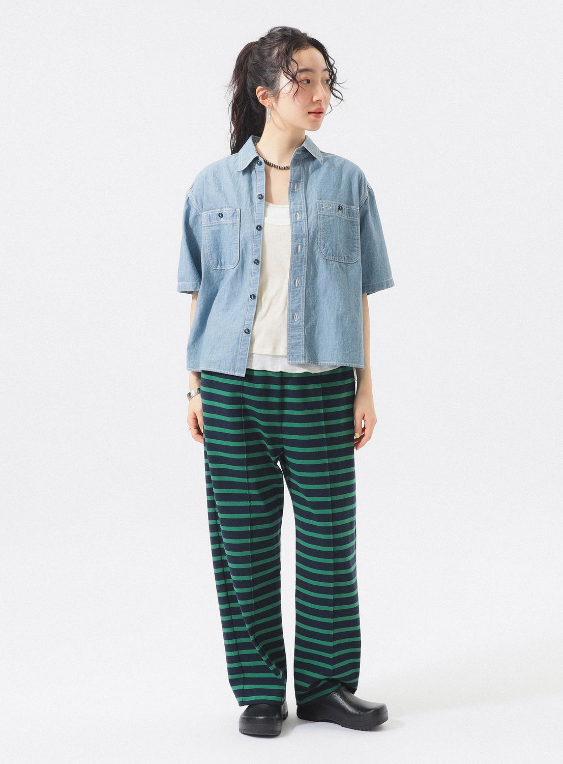 A woman stands against a white background wearing a BEAMS BOY Chambray Short-Sleeve Work Shirt over a white top, green and navy striped pants, black shoes, and a necklace. She looks to the side with her hands at her sides.