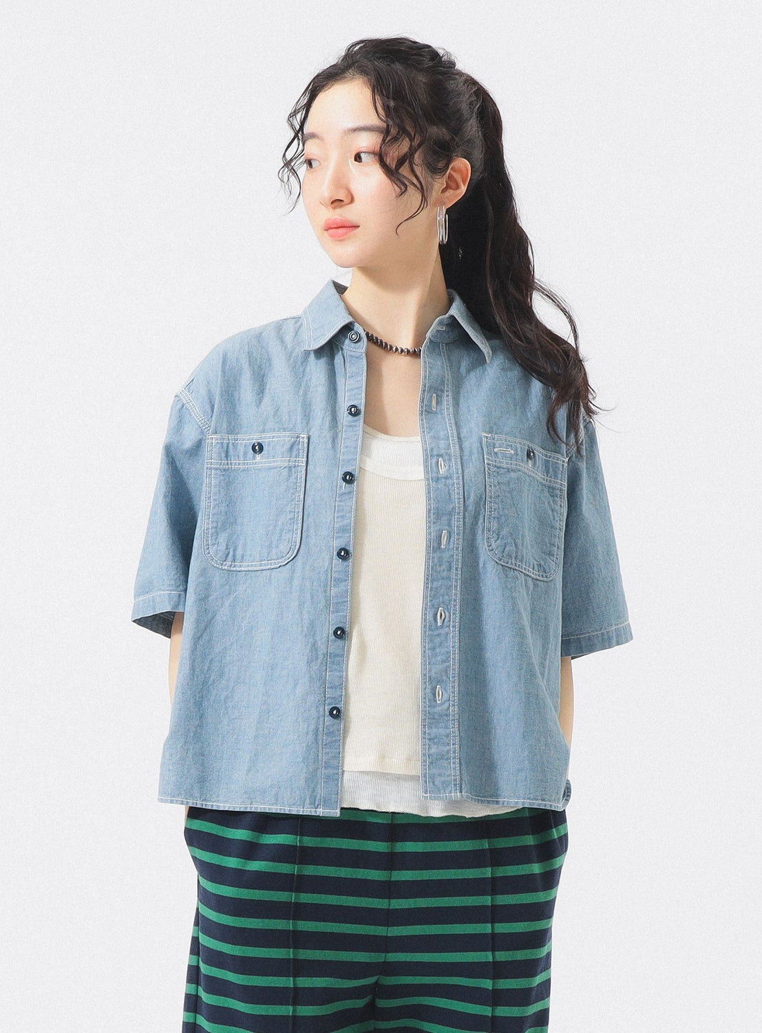 A person with long, wavy hair tied back wears a BEAMS BOY Chambray Short-Sleeve Work Shirt over a white top and green-striped pants, standing against a plain white background and looking to the side.