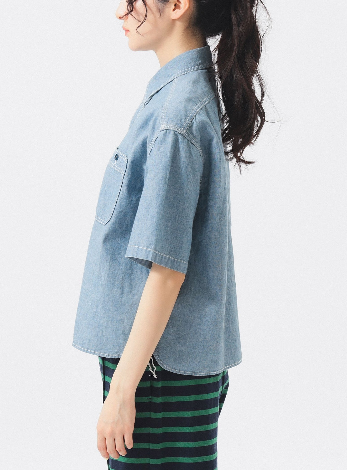 A person with long, wavy dark hair in a ponytail is seen in profile, wearing a Chambray Short-Sleeve Work Shirt by BEAMS BOY and green-and-black striped pants, standing against a plain white background.