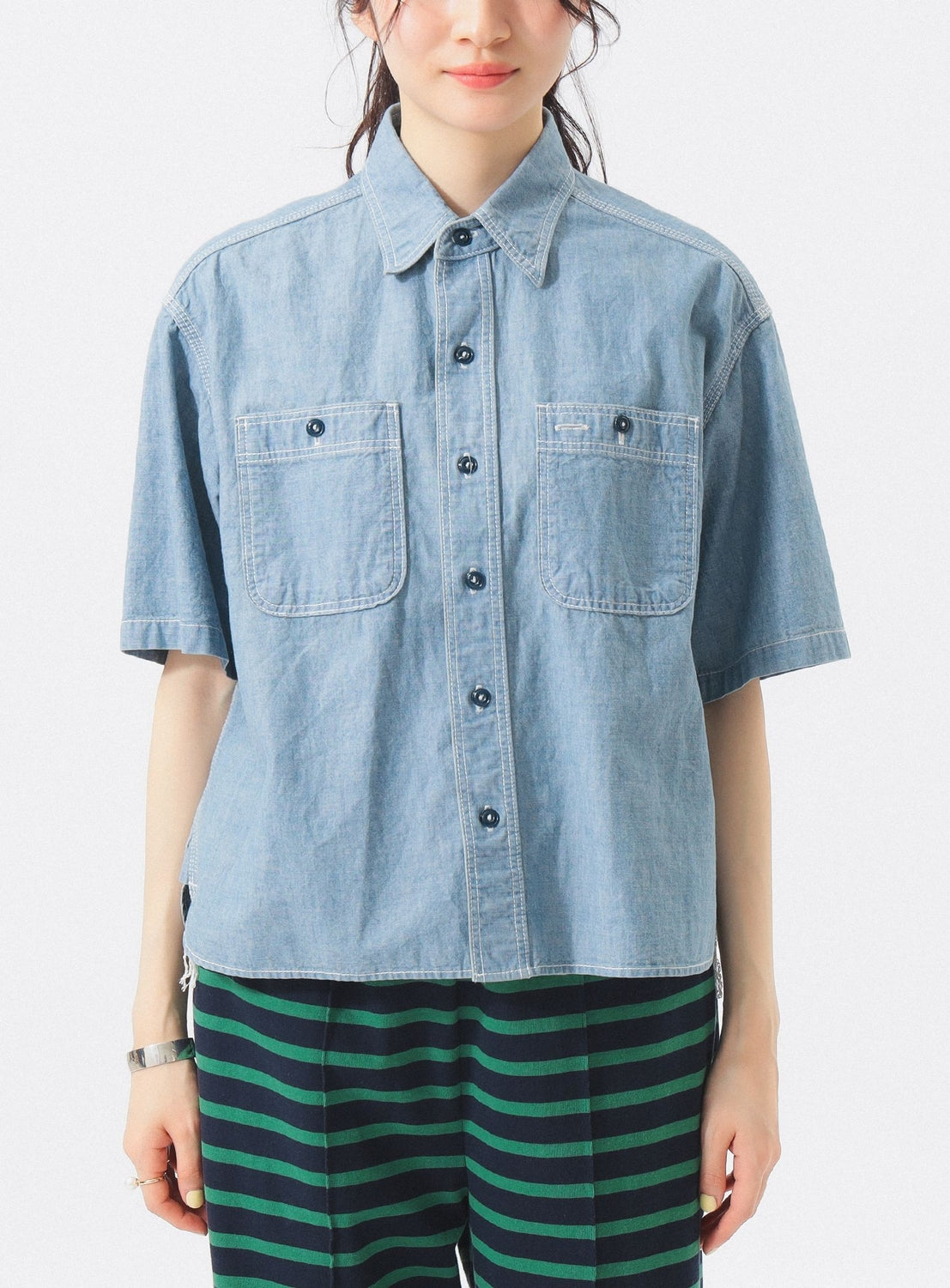 A person wears the BEAMS BOY Chambray Short-Sleeve Work Shirt with two chest pockets and relaxed fit, paired with green and black striped pants, standing against a plain white background.