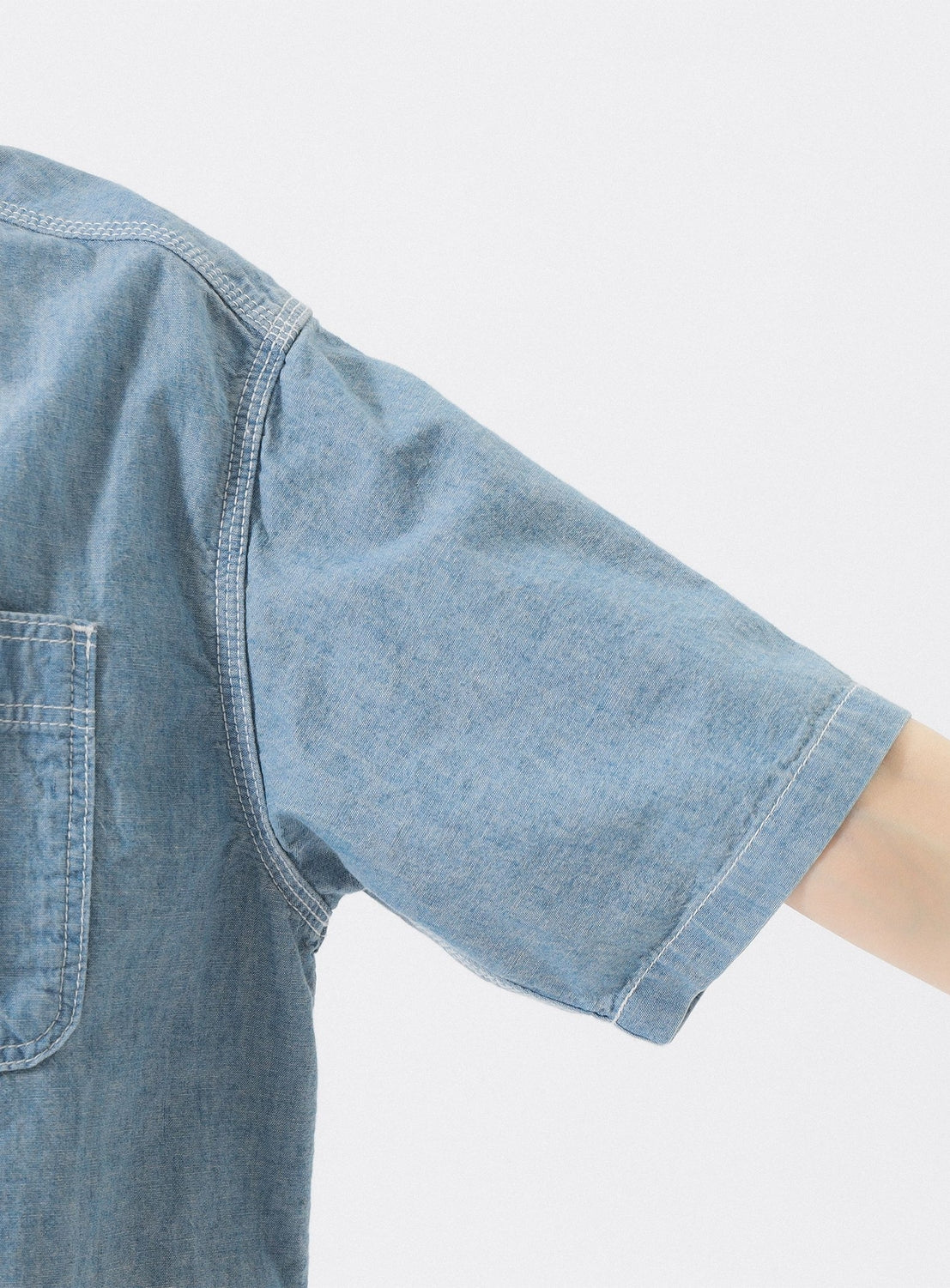 A close-up of an outstretched arm in the Chambray Short-Sleeve Work Shirt by BEAMS BOY, showing visible stitching and a partial pocket against a plain white background that highlights the shirt’s relaxed fit and light blue chambray fabric.
