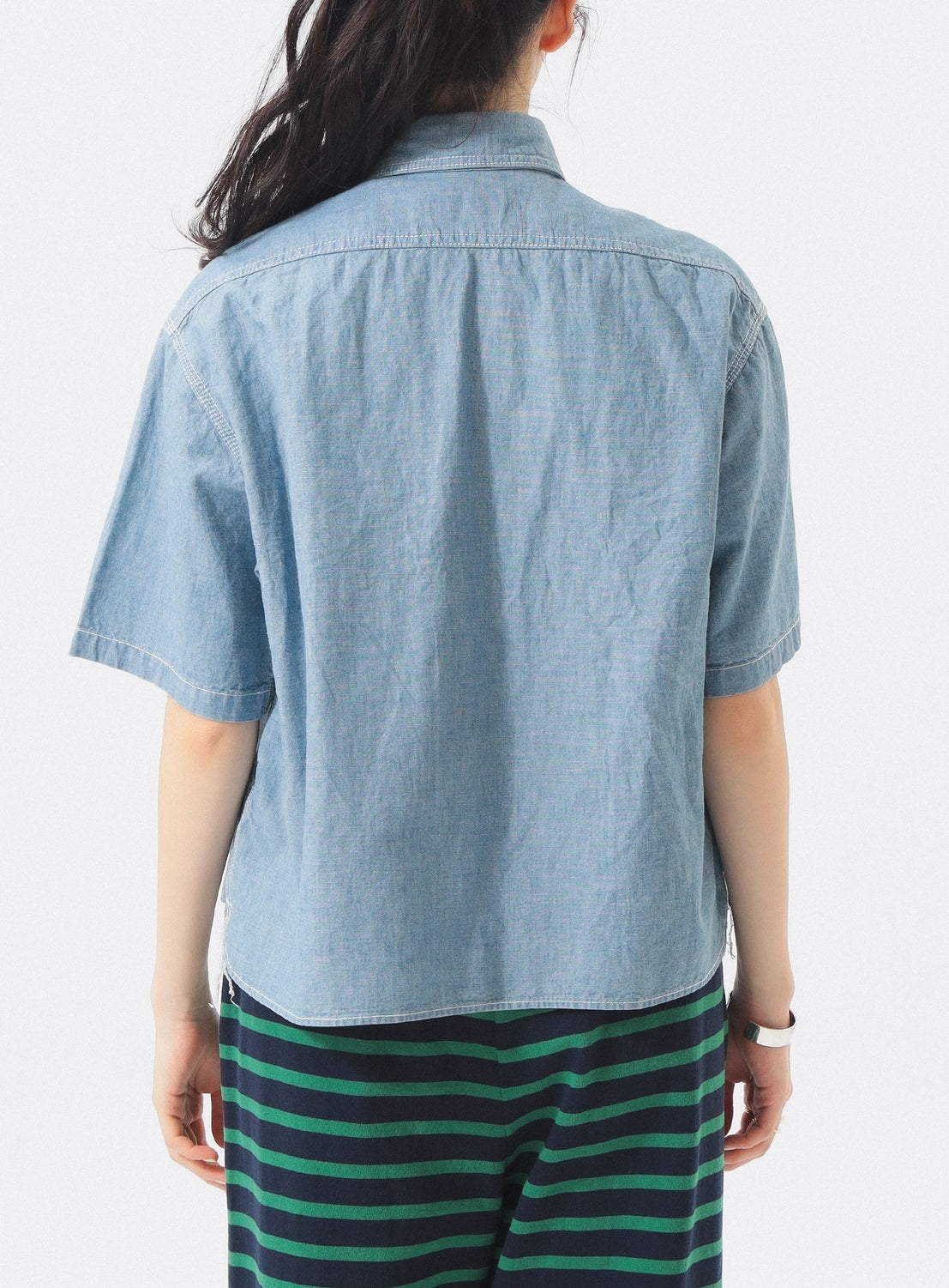 A person with long dark hair, wearing a BEAMS BOY Chambray Short-Sleeve Work Shirt and green-and-black striped pants, stands with their back to the camera against a white background.