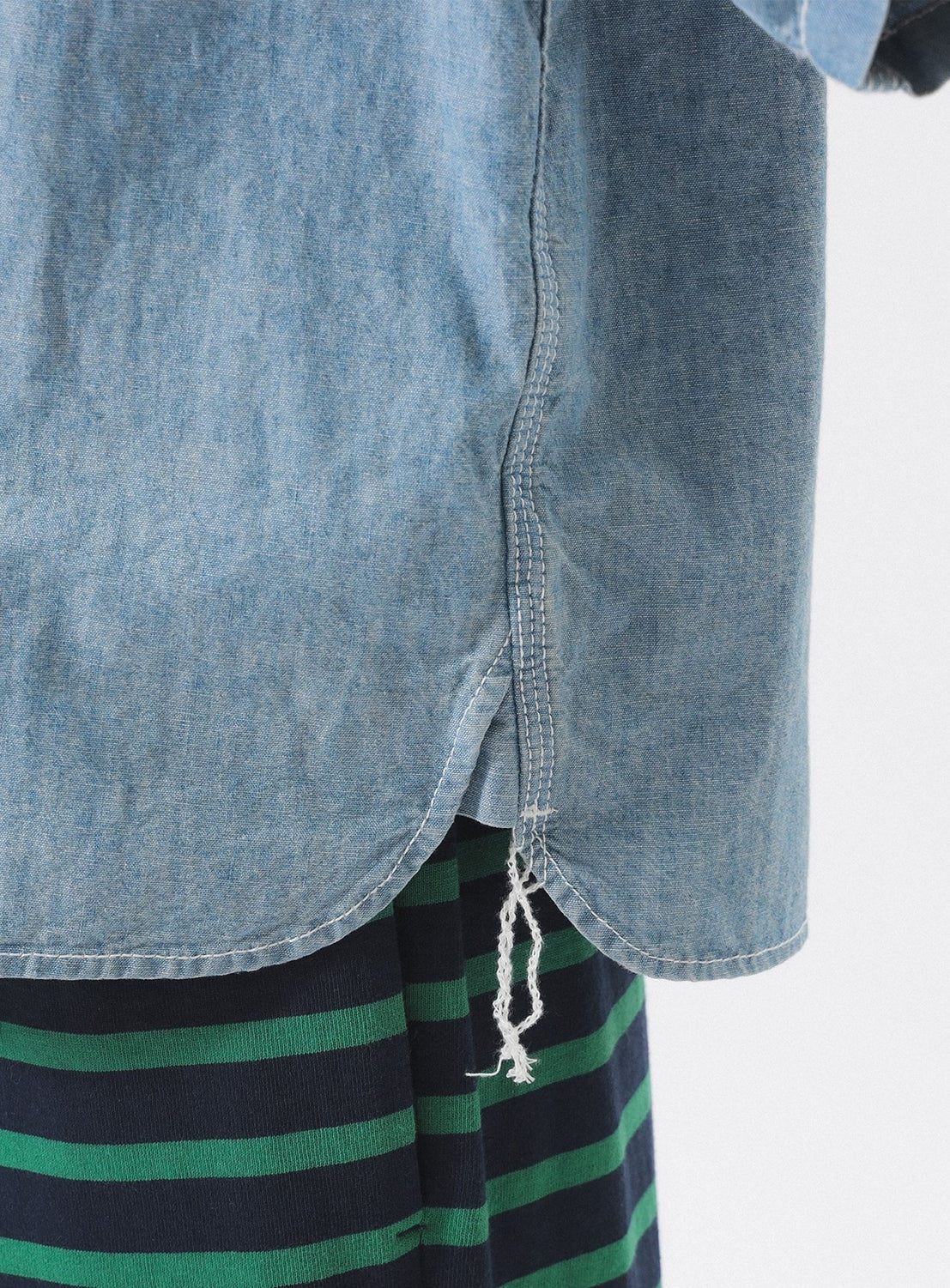 Close-up of the BEAMS BOY Chambray Short-Sleeve Work Shirt in light blue, showing a frayed seam and relaxed fit over a dark blue and green striped layer, set against a plain white background.