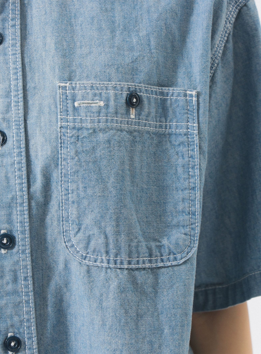 Close-up of the BEAMS BOY Chambray Short-Sleeve Work Shirt in blue, showing its relaxed fit, chest pocket with white stitching, black buttons, and part of the short sleeve and upper torso.