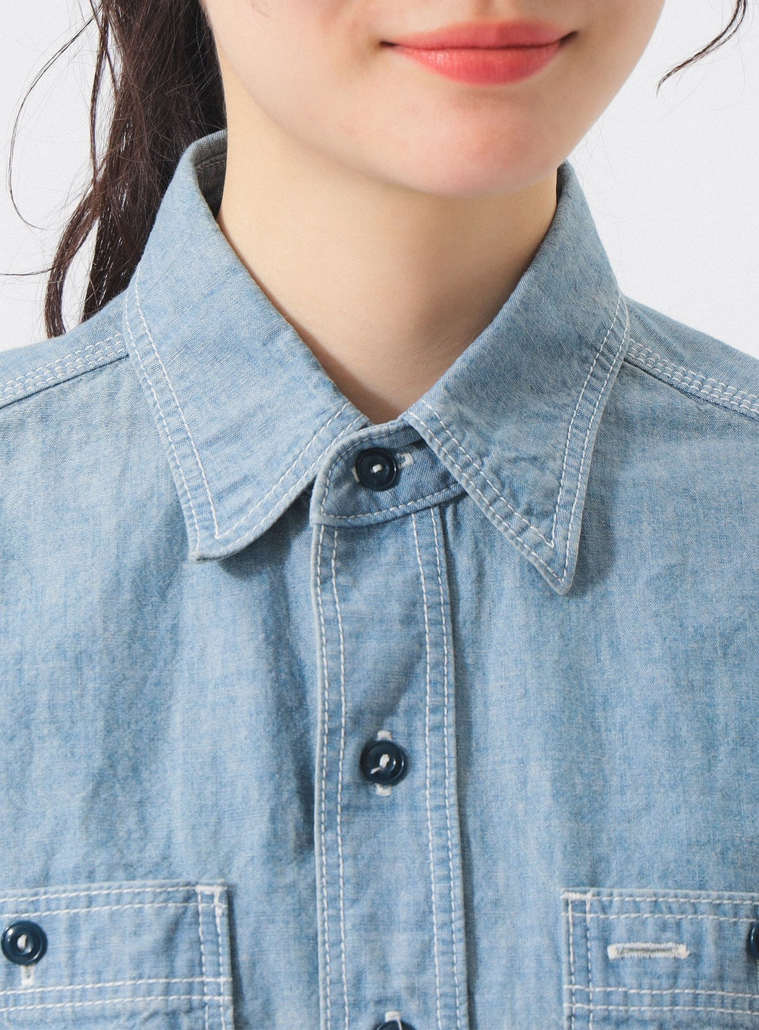 A person with light skin and dark hair wears the BEAMS BOY Chambray Short-Sleeve Work Shirt. Only the lower half of their face and upper torso are shown, emphasizing the shirt’s relaxed fit.
