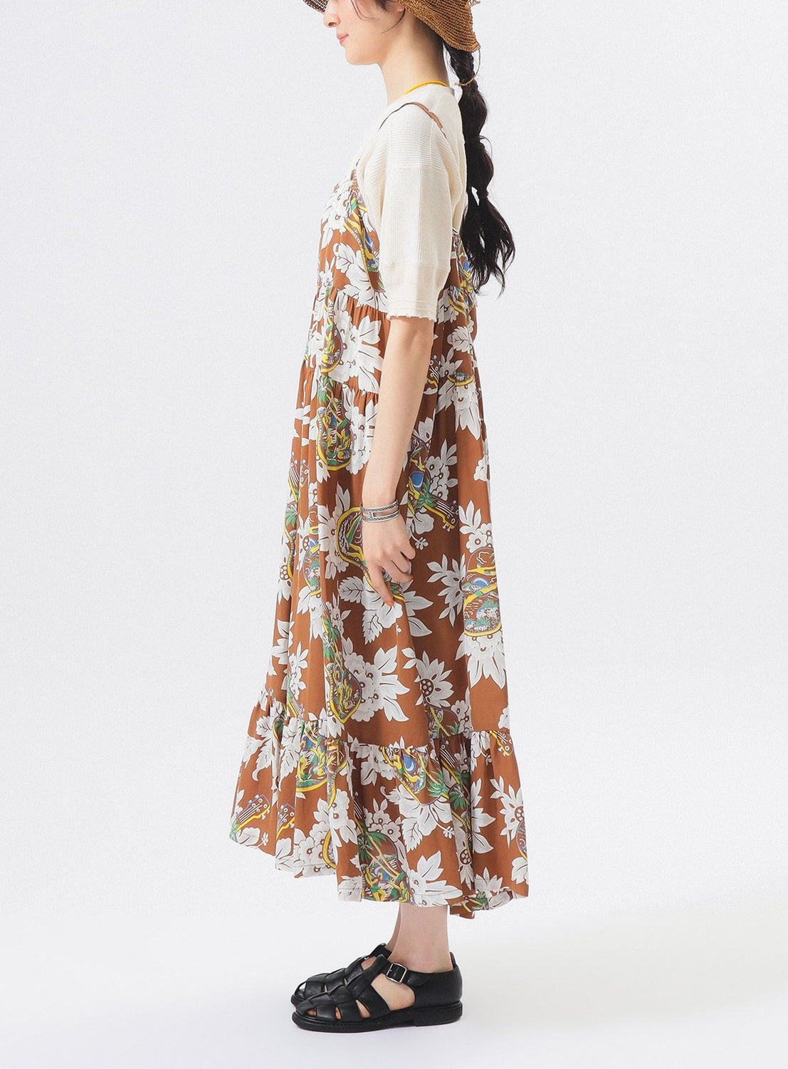 A woman in profile wears the [Exclusive] SUN SURF Sleeveless Cami Dress by SUN SURF over a light short-sleeve top, with black sandals and a brown hat, her long hair tied back, standing against a plain white background.