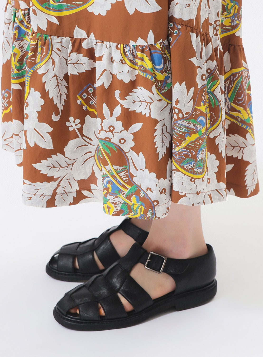 A person stands on a white surface wearing the SUN SURF [Exclusive] SUN SURF Sleeveless Cami Dress, black woven leather sandals, and a brown floral-paisley skirt, embodying relaxed resort style.