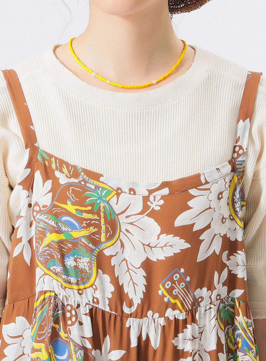 A person wears a cream ribbed long-sleeve top under the [Exclusive] SUN SURF Sleeveless Cami Dress by SUN SURF, featuring colorful floral designs. They accessorize with a yellow beaded necklace and a partially visible brown hat. Face not visible.