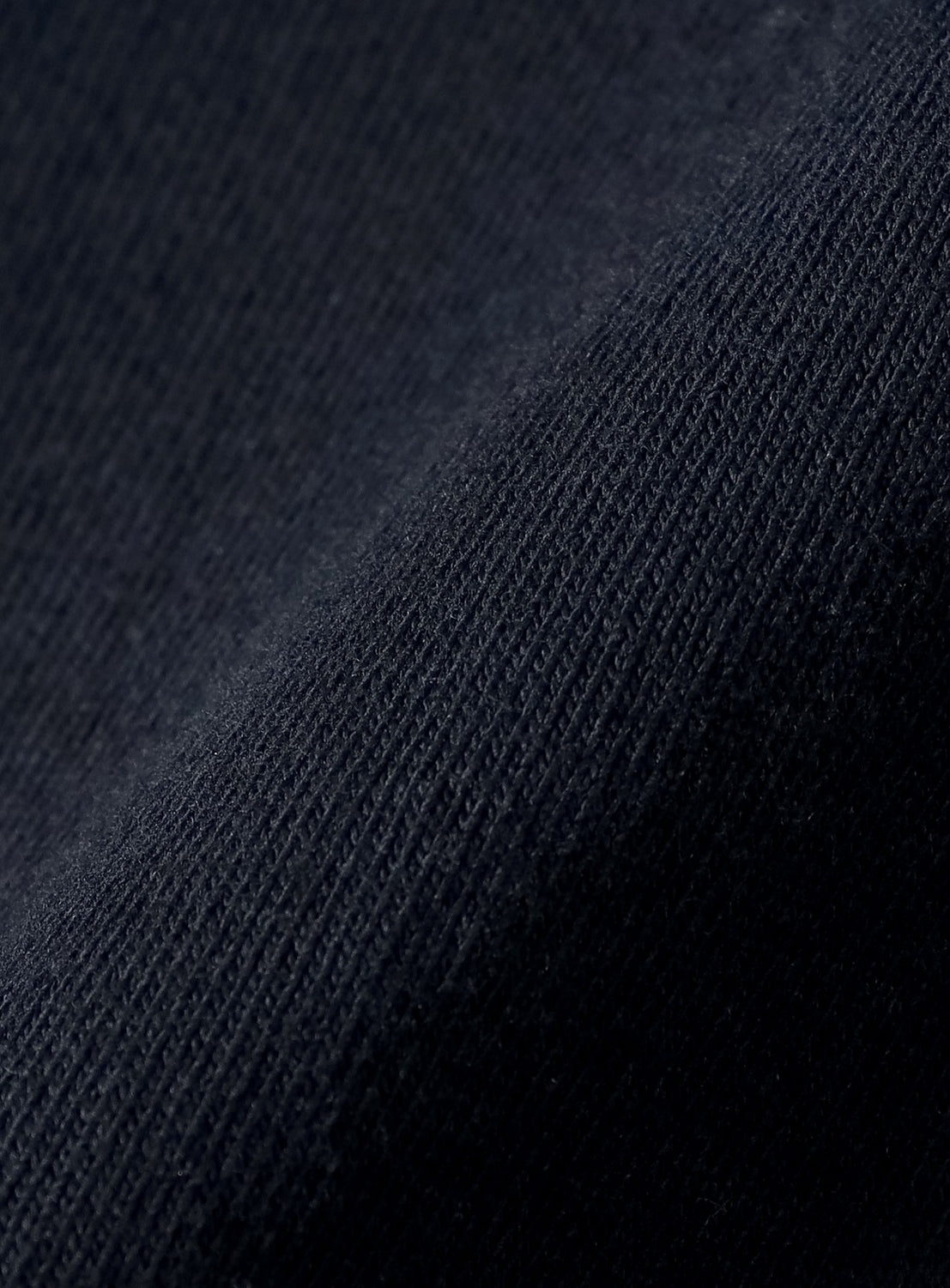 Close-up of a dark blue or black heavyweight US cotton fabric with a visible, textured weave pattern. The material is slightly wrinkled, revealing intricate detail in the fibers and thread lines—reflective of the premium quality featured in the BEAMS T Tyrrell Winston - W Logo T-Shirt.