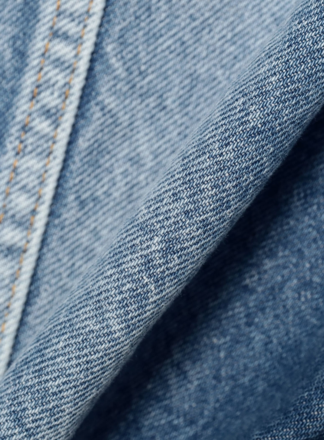 A close-up view of the INSCRIRE Used Collarless Denim Zip Jacket's heavyweight cotton denim fabric, highlighting the detailed weave texture and a seam accented with orange stitching.