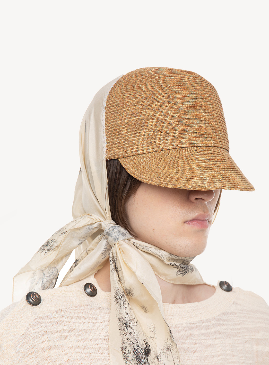 A person wears the DAIRIKU Scarf Docking Straw Cap, featuring a brown textured straw cap seamlessly combined with a cream silk scarf adorned with a black floral pattern and tied around their head. The elegant Japanese-made scarf adds sophistication, while their face is partially obscured by the cap. They are dressed in a cream top with button details on the shoulders.