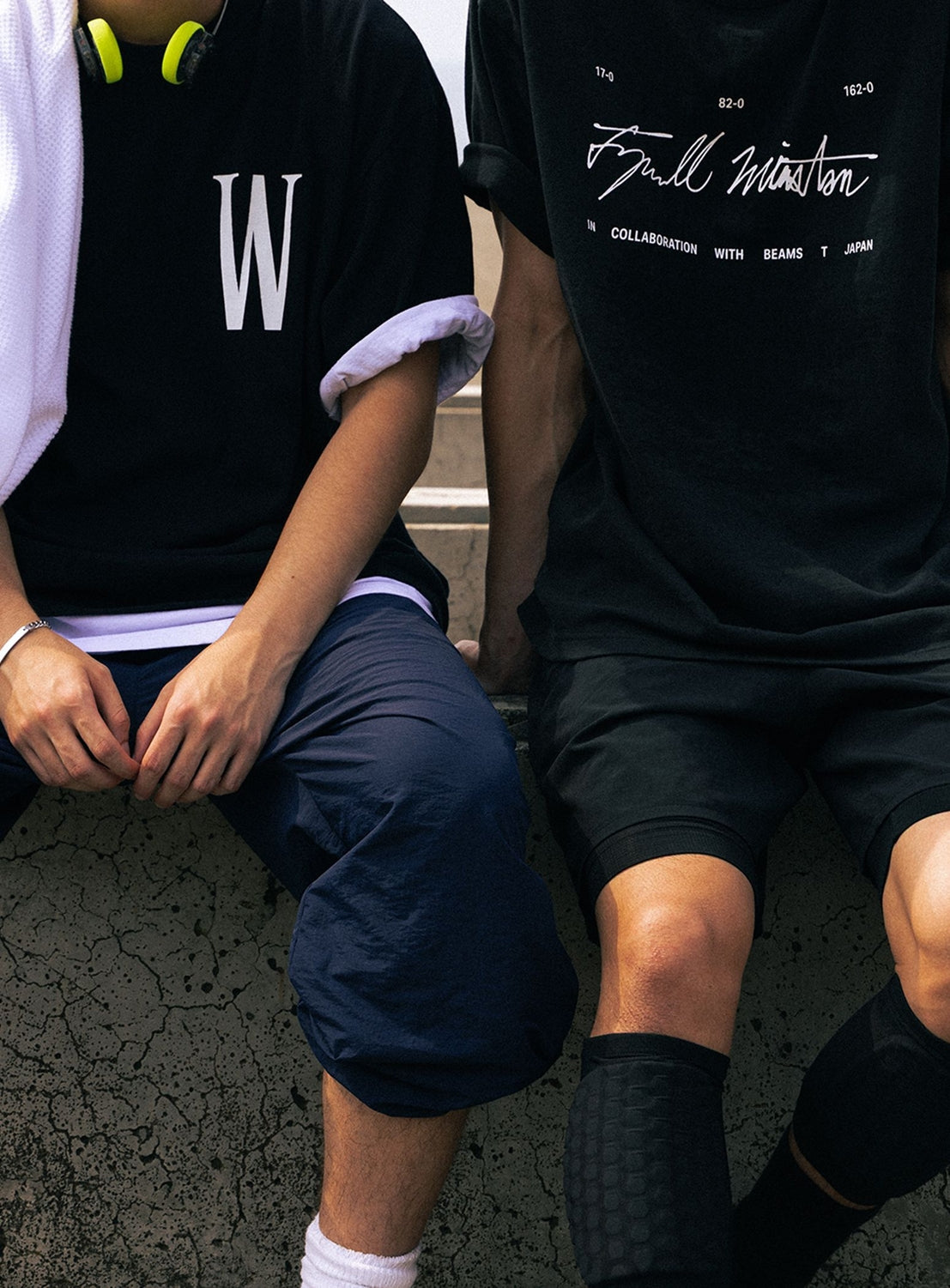 Two people sit side by side outdoors, both in black T-shirts—one wears the BEAMS T Tyrrell Winston - W Logo T-Shirt, crafted from heavyweight US cotton and featuring a bold white ‘W’ logo; the other sports a distinct design inspired by Tyrrell Winston with unique white text. Their faces are not visible. One person has headphones around their neck and a towel draped over their shoulder.