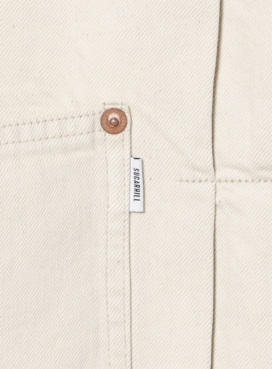 Close-up of an off-white SUGARHILL Tattoo Flash Print Denim Jacket in premium denim, showing visible stitching, a metal rivet, and a small white SUPRAVIVAL tag by the pocket.