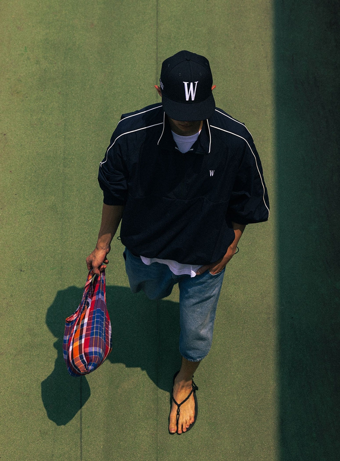 A person dressed in the BEAMS T Tyrrell Winston Anorak Jacket, denim shorts, a black cap and shirt, and flip-flops walks across a green surface while carrying a colorful plaid bag. The overhead shot features their shadow on the right side, subtly echoing vintage athletic style reminiscent of WIN SPORT branding.