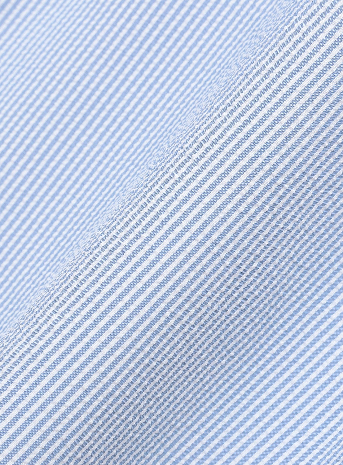 Close-up of the light blue and white striped fabric from the BEAMS Stretch Quick-Dry Seersucker Relaxed Fit Shirt, highlighting its lightweight, wrinkle-resistant texture with fine diagonal lines. The seersucker weave creates a soft, repetitive pattern with gentle folds visible across the surface.