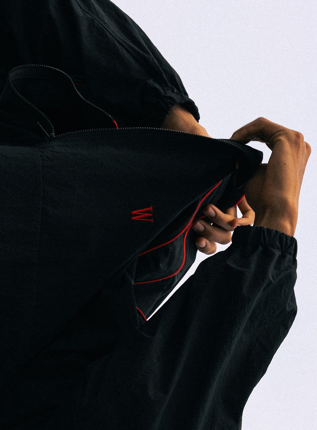 Wearing the BEAMS T Tyrrell Winston - Anorak Jacket, a person zips up the vintage-inspired black outerwear with red lining, concealing part of their face. Only their hands and arms are visible against a simple light background.