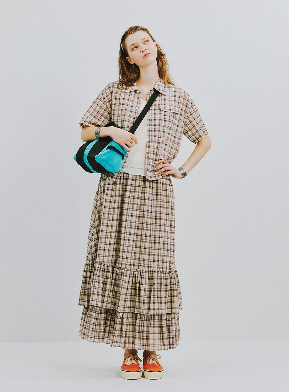 Wearing a BEAMS BOY Check Tiered Skirt and a matching check-pattern short-sleeve shirt made from lightweight fabric, along with a white top, orange sneakers, a headband, and carrying a blue bag over their shoulder, the person stands against a plain light background.
