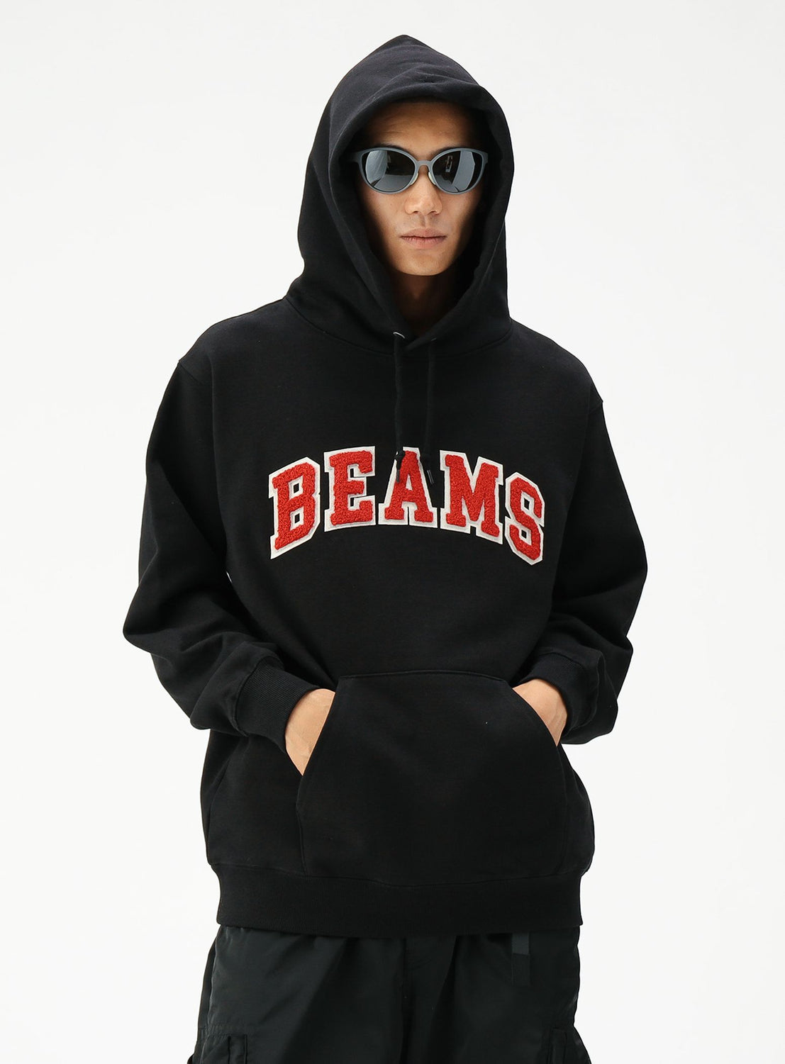 A person wears a black BEAMS Basic Hoodie with red and white chenille patches, black sunglasses, and the hood up, standing against a plain white background. The BEAMS Basic Hoodie offers a comfortable fit for everyday wear.