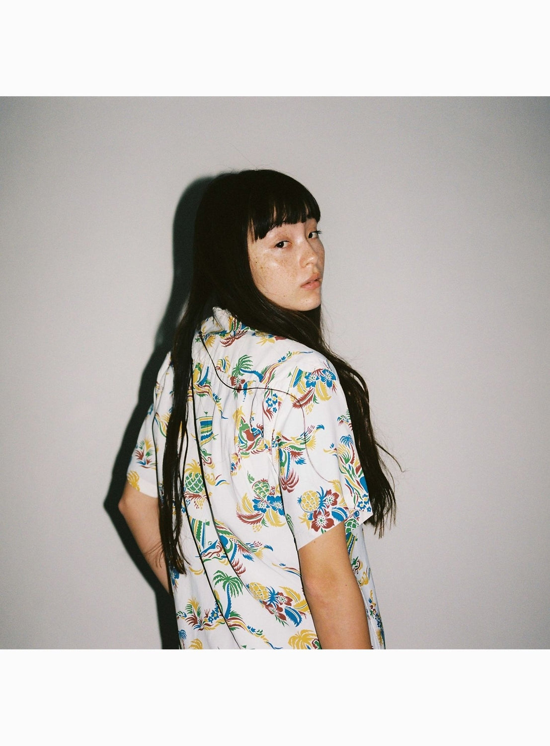 A person with long dark hair and bangs, turned slightly away and looking at the camera, wears the [Exclusive] SUN SURF x BEAMS BOY Open Collar Shirt by SUN SURF—a short-sleeved white shirt with a playful Hawaiian floral and bird pattern.
