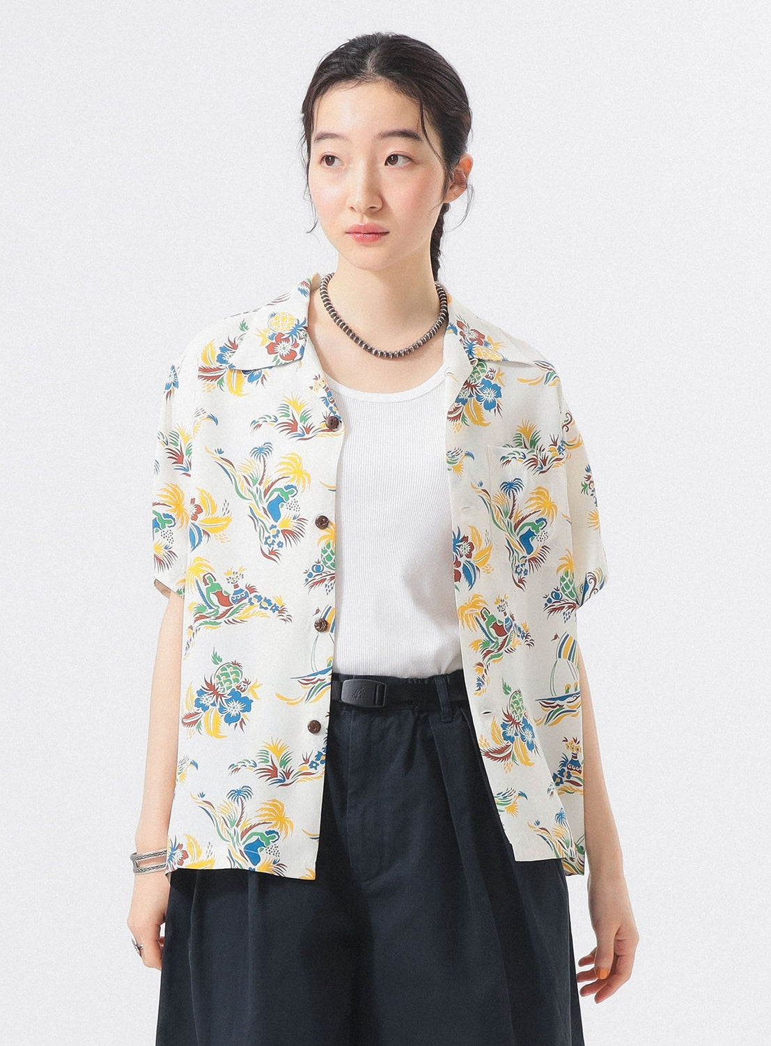 A woman with dark hair tied back models the [Exclusive] SUN SURF x BEAMS BOY Open Collar Shirt by SUN SURF over a white top and dark pants, accessorized with a necklace and bracelet, standing against a plain white background.